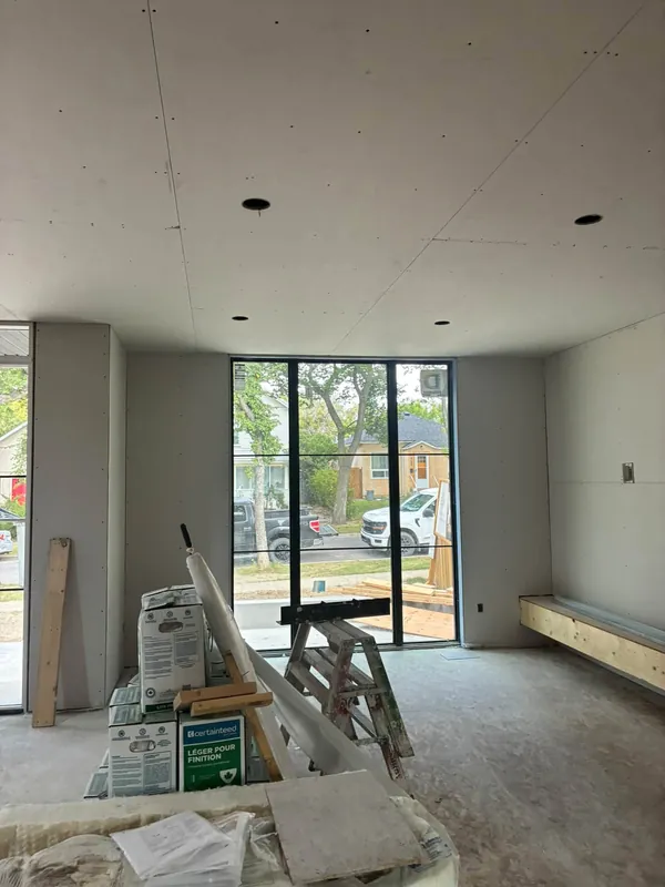 A room with large sliding glass doors, newly boarded walls and ceiling, with a ladder and materials staged on the floor